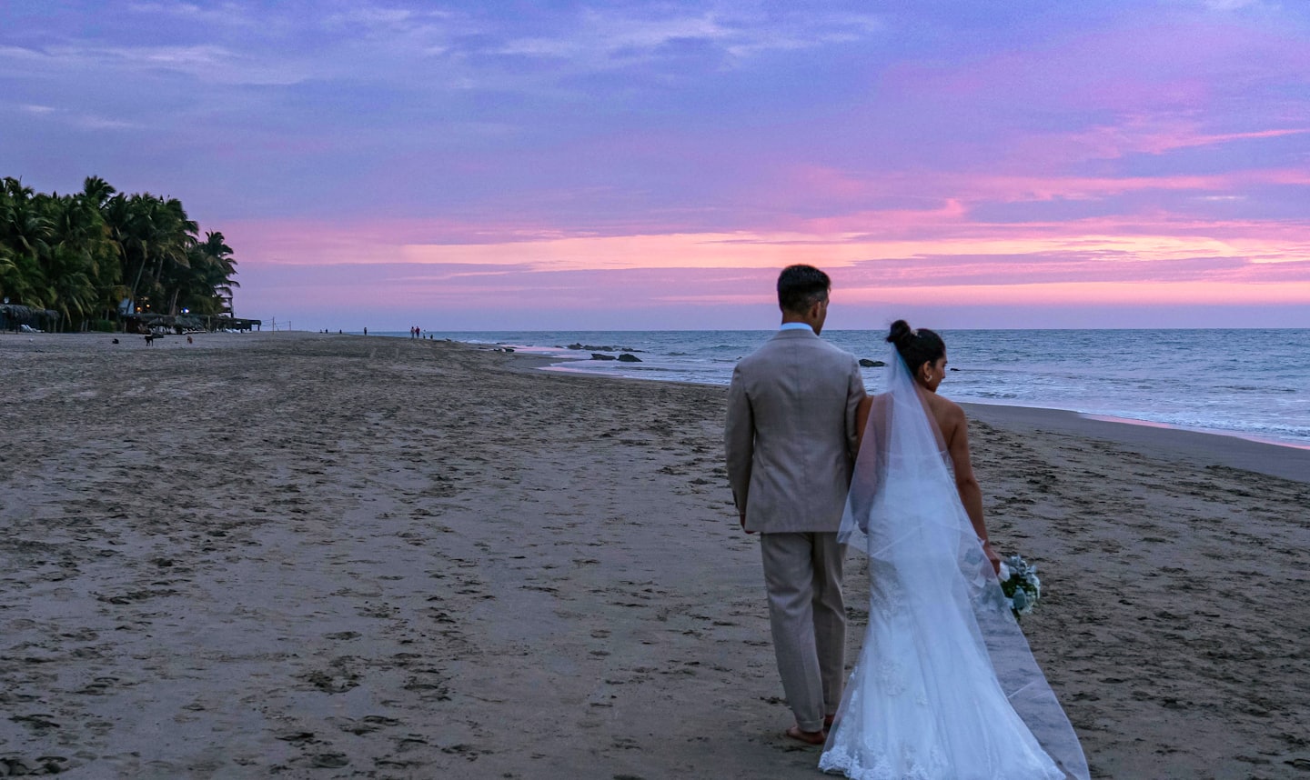 Recién casados caminan por la playa al atardecer con palmeras.